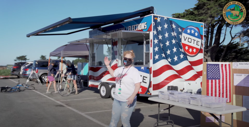 A woman stands in front of a mobile trailer designed with American flags and "VOTE" stickers. The VoteMobile helps increase accessibility in elections.