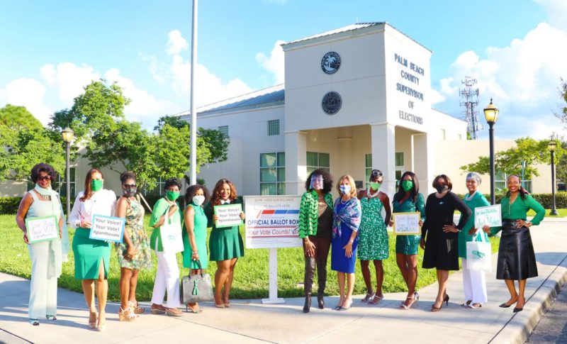 A group of women dressed in formal attire pose by a ballot box.