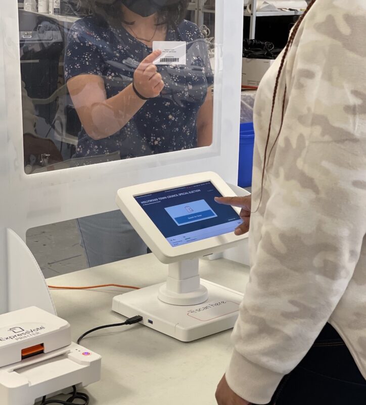 A voter holds up their driver's license behind a plastic screen while another person clicks a touch screen.