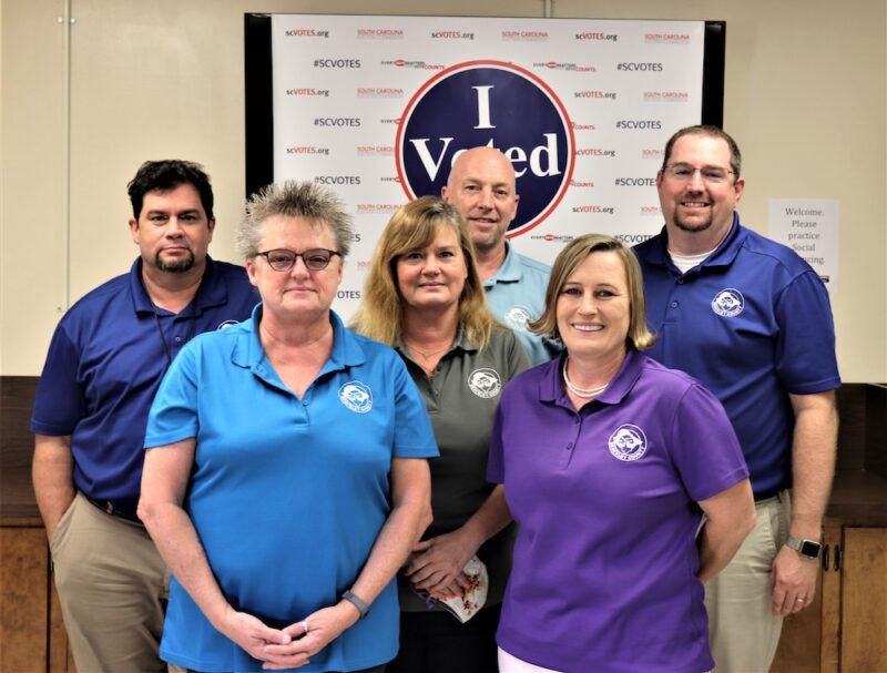 Six adults pose for a photo in front of an "I Voted" banner