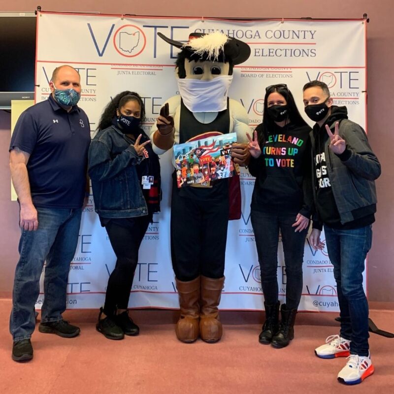 Four people wearing masks pose with a mascot in front of a sign that says "Cuyahoga County Board of Elections"