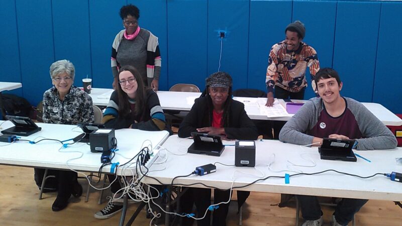 Six adults sit and stand behind tables with election equipment. 