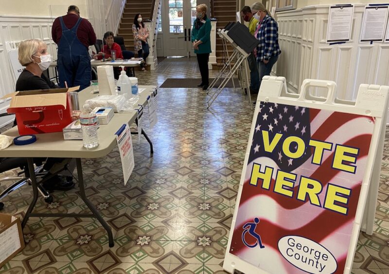 A "Vote Here" sign is in the foreground of the photo. In the background, election workers sit at tables and voters stand by voting machines.
