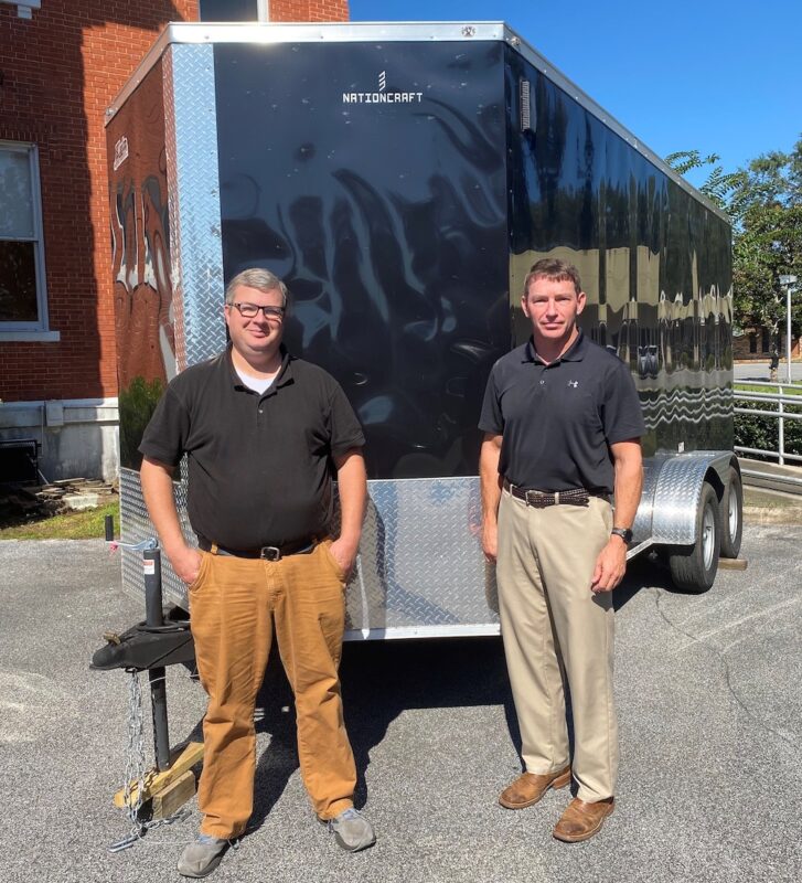 Two men stand in front of a large trailer.