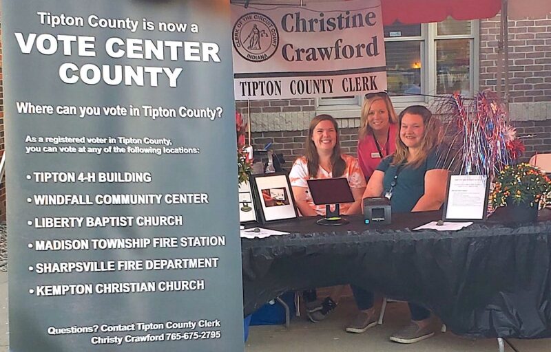 Three women from the study committee sit at a booth with an electronic poll book and a sign that says "Tipton County is a Vote Center County"