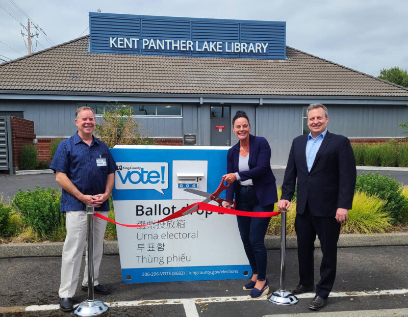 three people pose in front of the library with a ballot drop box