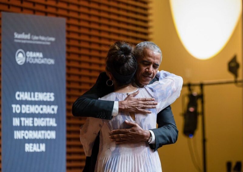 Former President Barack Obama embraces a woman on a stage. In the background is a sign that says "Challenges to democracy in the digital information realm."
