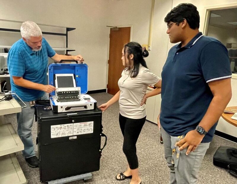 A man shows a machine to two younger people.