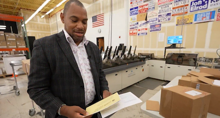 A man holding an absentee ballot poses with a machine and computer behind him.