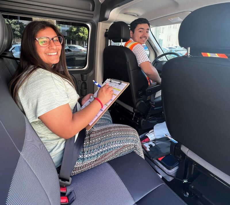 a woman in the back seat of a vehicle with a clipboard and a man in the driver seat smile