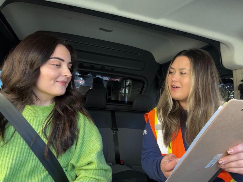 two women inside a vehicle. one woman shows a clipboard to the other.